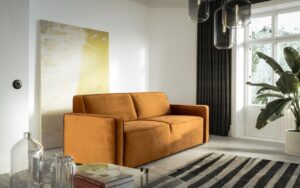 Złożna sofa Muto w żółtej tapicerce stoi w salonie pod ścianą z wielkim obrazem.