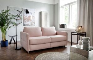 Mała złożona sofa Ema w jasnej różowej pastelowej tapicerce stoi w salonie na tle ściany z obrazem i kaloryferem.